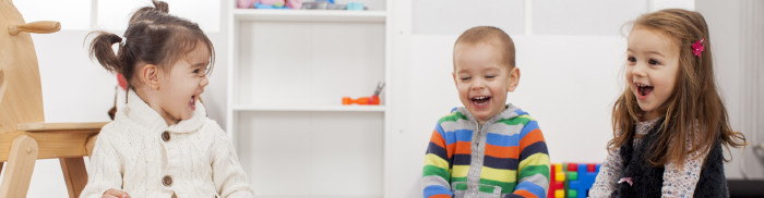 © Boggy, stock.adobe.com Zwei kleine Mädchen und ein Bub sitzen im Kindergarten und lachen